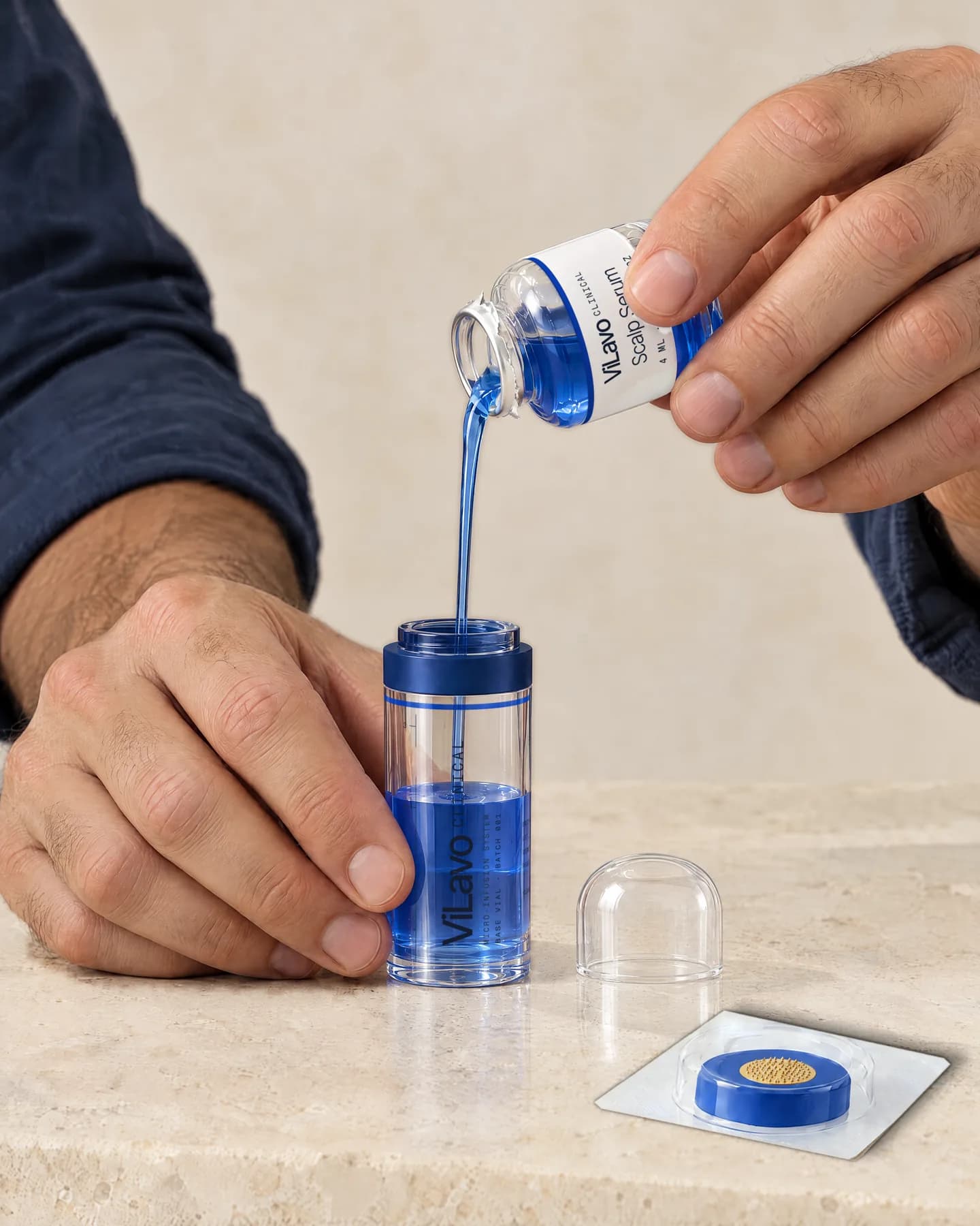 A man pours a small vial of blue serum into the clear base vial, its dome cap set aside on the stone.